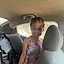 braided_hair, car_interior, car_seat, casual_clothing, child, child_seat, daylight, dress, fruit_pattern, happy, indoor, person, portrait, seat_headrest, seatbelt, smiling, sunlight, vehicle, window, young_child