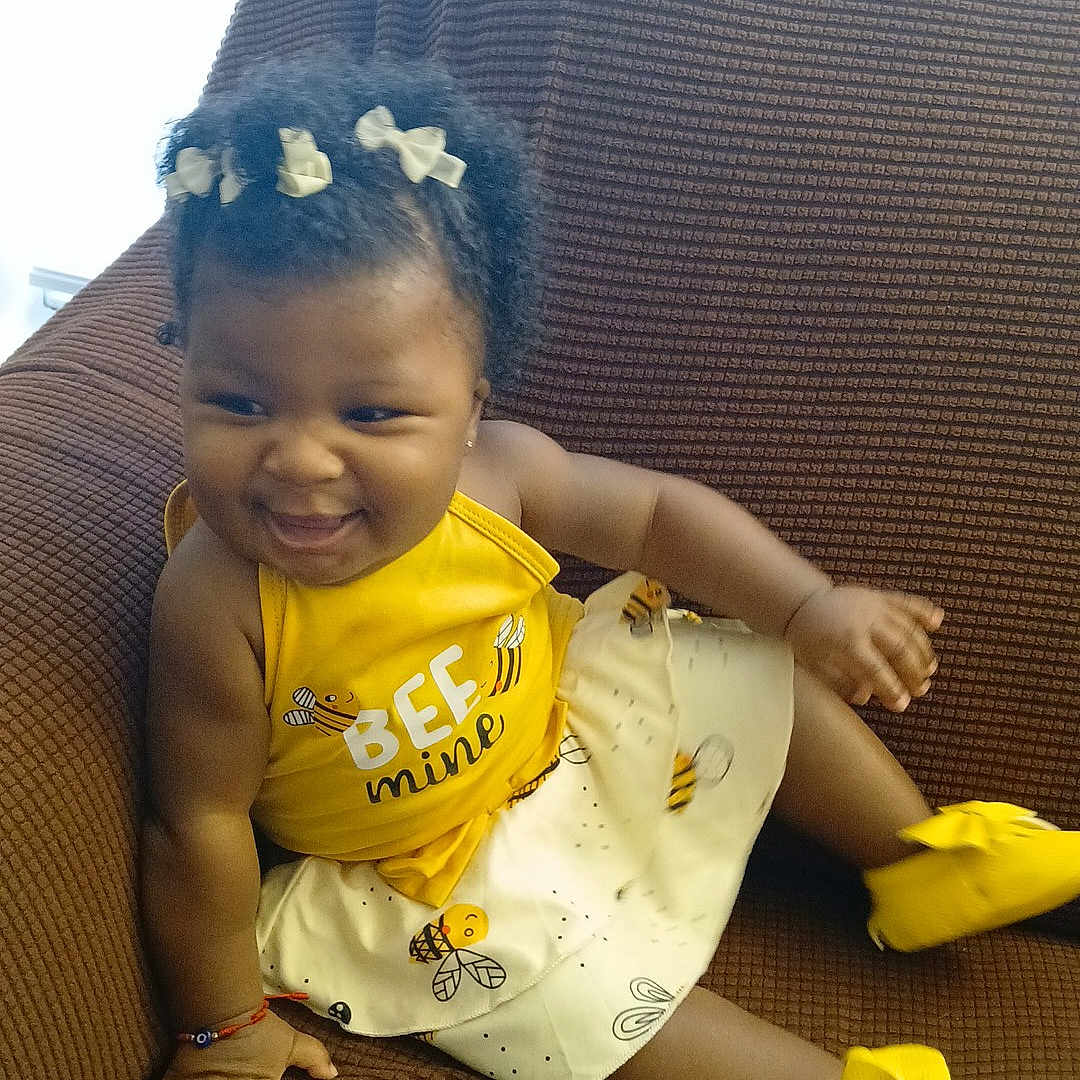 Nanyamka is registered to the contest to win money with this photo: accessory, baby, brown_couch, child, couch, curly_hair, cute, dress, fashion, footwear, hair_bows, happy, indoors, person, playful, portrait, sitting, smiling, toddler, yellow_clothing