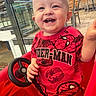 toddler, child, smiling, red_shirt, spiderman, toy_car, steering_wheel, indoor, restaurant, chairs, tables, happy, person, casual, playful, baby, seatbelt, shorts, bright_colors, cheerful