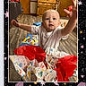 baby, child, indoor, wooden_floor, toy, wrapping_paper, white_shirt, reaching_hand, curious_expression, cabinet, door, flooring, digital_frame, stars, moon, social_media_interface, reaction_icons, colorful, celebration, portrait
