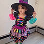 child, costume, witch_hat, colorful_clothing, polka_dots, rainbow_stripes, boots, indoor, floor, wooden_floor, curly_hair, long_sleeves, black_hat, purple_glitter, dress, tights, standing, person, cute, playful