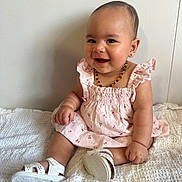 Mahlani joined the competition — help win amazing prizes! baby, child, smiling, sitting, pink_dress, white_sandals, beaded_necklace, earrings, indoor, blanket, happy, cute, portrait, infant, young_child, fashion, accessories, footwear, person, wall