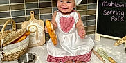 Mahlani is registered to the contest to win money with this photo: baby, child, chef_hat, apron, bread, baguette, kitchen, flour, baking_tools, whisk, rolling_pin, basket, cutting_board, chalkboard, smiling, sitting, tile_wall, countertop, messy, happy