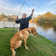 Hunter is registered to the contest to win money with this photo: person, dog, fish, lake, grass, fishing_rod, water, trees, autumn, outdoor, animal, pet, catch, nature, recreation, happy, sitting, casual_clothing, collar, sky
