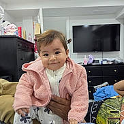 Ahyse is registered to the contest to win money with this photo: adult_hand, baby, blanket, child, couch, dresser, flower_pattern, hair_bows, happy, home, indoor, living_room, pink_jacket, plush_toy, portrait, smiling, standing, television, toddler, white_outfit