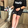 cat, person, indoor, wooden_floor, tile_wall, black_clothing, glasses, shorts, socks, petting, curious_cat, tattoo, smartwatch, counter, sitting, standing_cat, light_skin, short_hair, casual, home