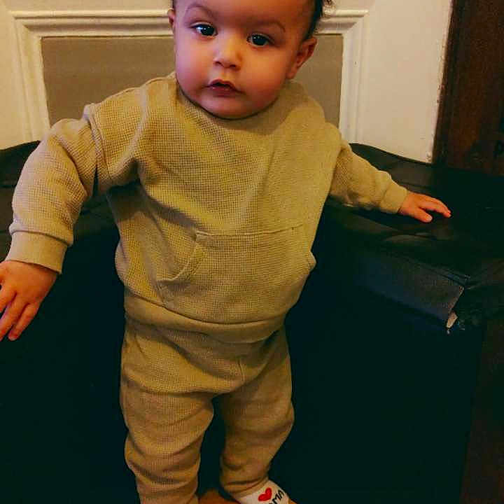 Naïm a rejoint le concours — aidez-le/la à gagner de superbes lots ! toddler, child, indoor, standing, clothing, socks, wooden_floor, cushion, black_furniture, face, curly_hair, sweatshirt, pants, expression, home, portrait, young_child, flooring, casual_wear, day_of_week_text