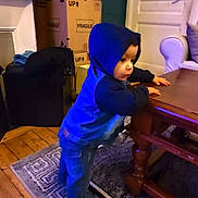 Naïm participe au concours pour gagner de l'argent avec cette photo : toddler, child, hoodie, blue_clothing, indoor, table, wooden_table, rug, carpet, furniture, boxes, fragile_label, floor, curious, standing, leaning, person, home, cozy, casual