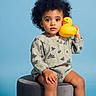 toddler, child, curly_hair, rubber_duck, stool, blue_background, casual_clothing, sneakers, sitting, portrait, cute, indoor, playful, young_child, person, studio, toy, expression, feet, fashion