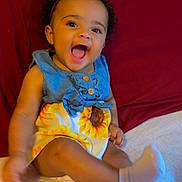 Camryn is registered to the contest to win money with this photo: baby, child, smiling, curly_hair, dress, sunflower, socks, bed, pillow, headboard, happy, sitting, indoors, person, cute, joyful, baby_girl, apparel, furniture, portrait