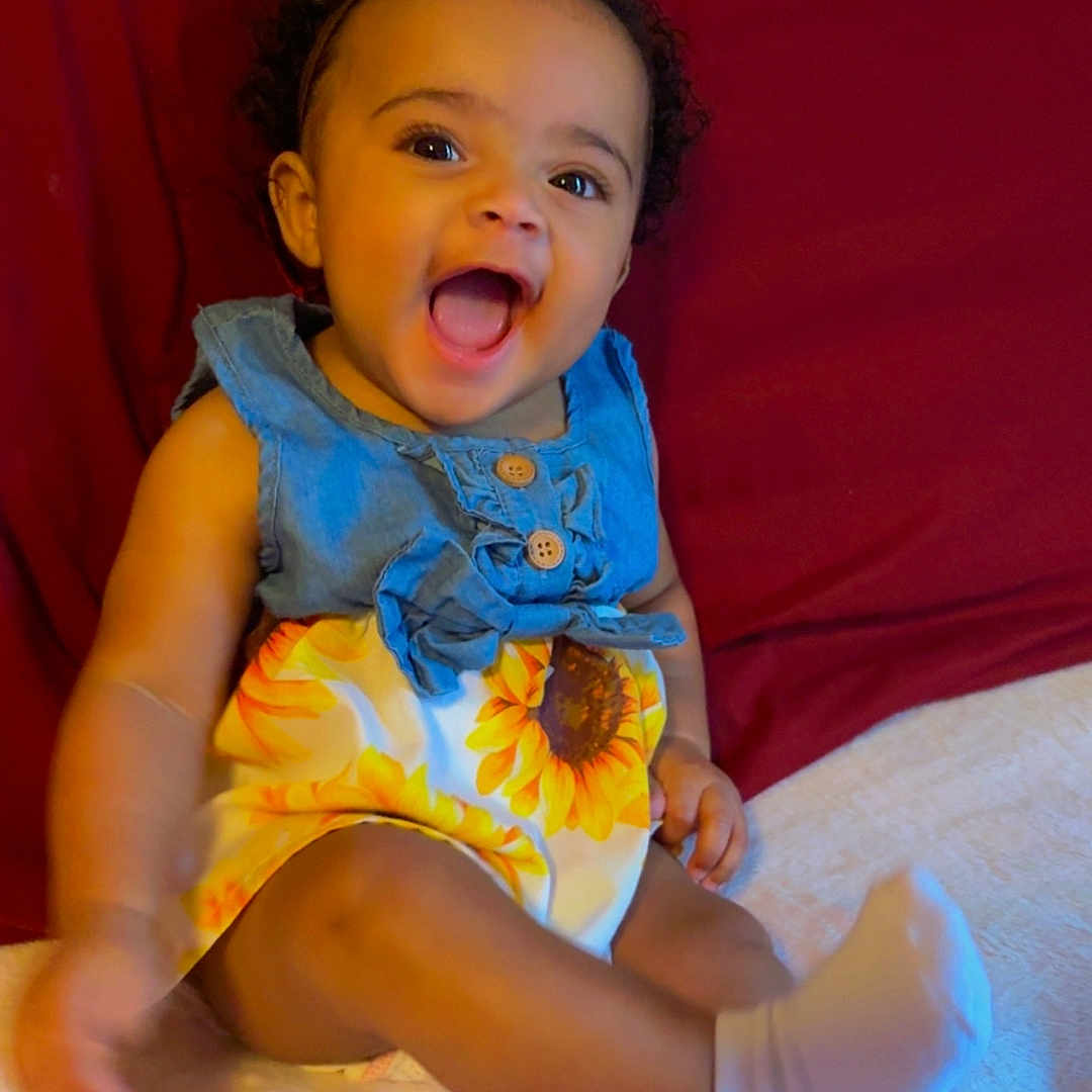 Camryn is registered to the contest to win money with this photo: baby, child, smiling, curly_hair, dress, sunflower, socks, bed, pillow, headboard, happy, sitting, indoors, person, cute, joyful, baby_girl, apparel, furniture, portrait