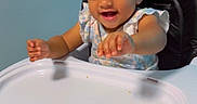 Camryn is registered to the contest to win money with this photo: baby, child, clothing, curly_hair, cute, face, footwearless, furniture, hair_beads, happy, high_chair, indoor, living_room, person, playful, reaching, smiling, stroller, table, toddler