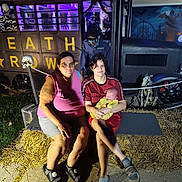 Aaron joined the competition — help win amazing prizes! woman, child, baby, hay_bale, halloween, decoration, skeleton, costume, night, outdoor, purple_light, vehicle, spooky, family, grass, concrete, person, glasses, shorts, tshirt