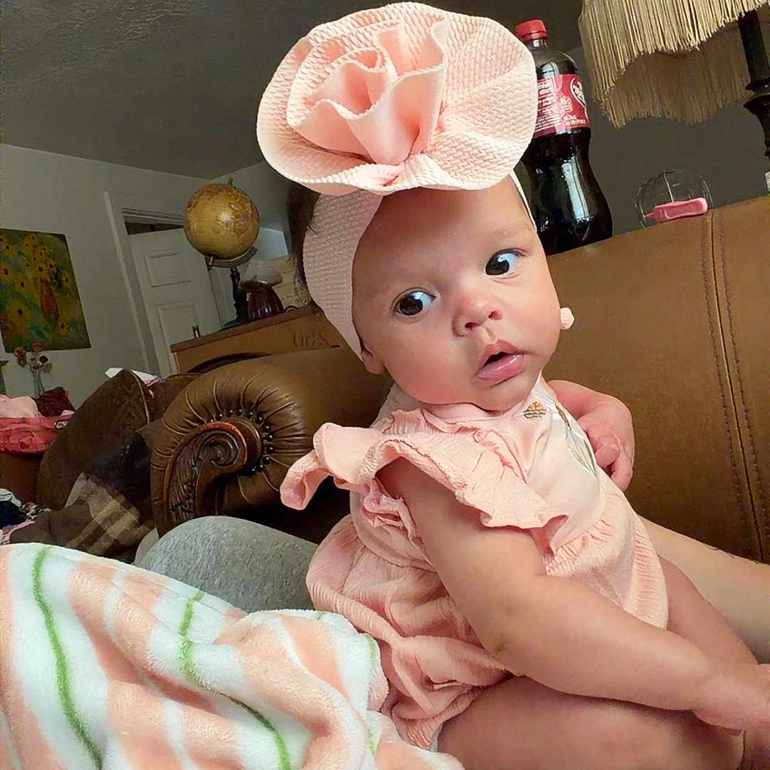 Royalynn joined the competition — help win amazing prizes! baby, infant, child, headband, flower, pink, dress, blanket, striped, couch, living_room, lamp, bottle, globe, furniture, curious, wide_eyes, seated, indoors, person