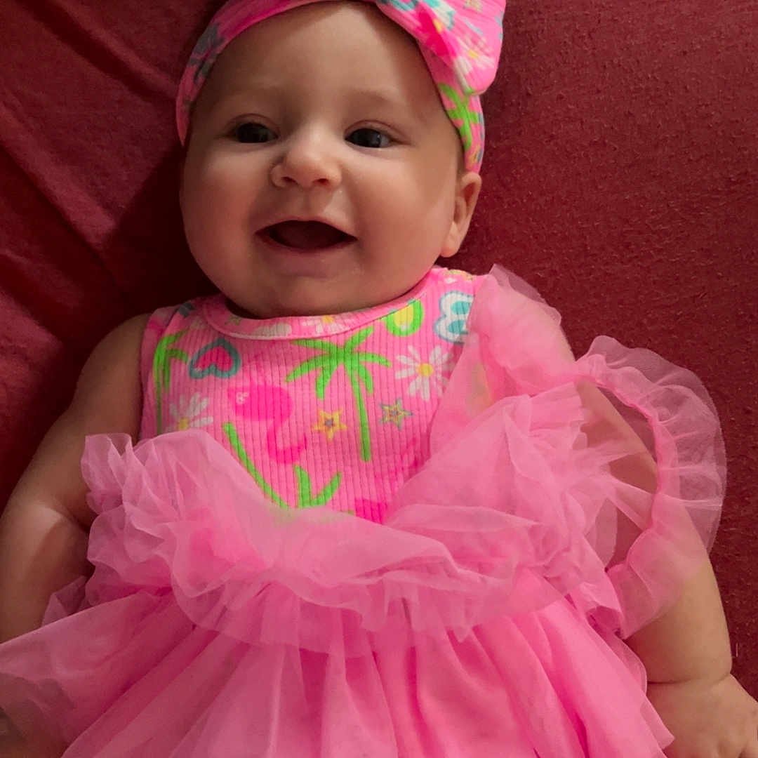 Addison is registered to the contest to win money with this photo: baby, blanket, bow, cheerful, child, cute, fabric, face, happy, headband, indoors, infant, laying_down, person, pink_dress, playful, portrait, skin, smiling, tutu
