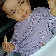 Josephine is registered to the contest to win money with this photo: toddler, child, high_chair, purple_sweater, unicorn_pattern, food, snack, indoor, person, face, hand, curly_hair, baby, expression, seated, cute, cozy, clothing, portrait, small_child