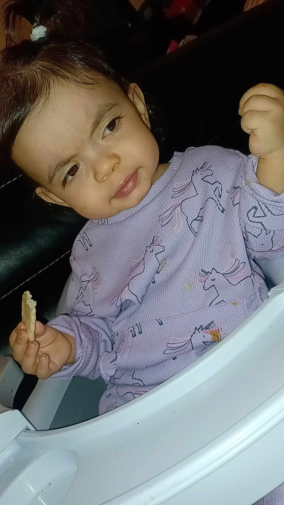 Josephine is registered to the contest to win money with this photo: toddler, child, high_chair, purple_sweater, unicorn_pattern, food, snack, indoor, person, face, hand, curly_hair, baby, expression, seated, cute, cozy, clothing, portrait, small_child