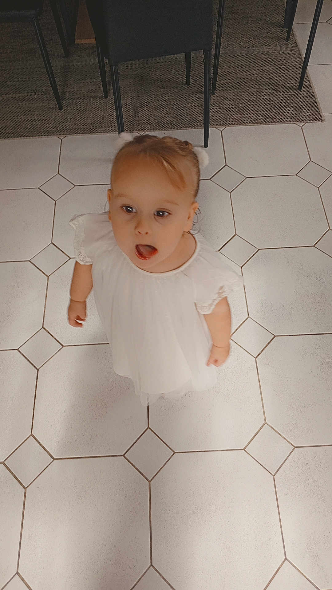 Livia participe au concours pour gagner de l'argent avec cette photo : child, toddler, white_dress, floor, tiles, pattern, indoor, curious, looking_up, standing, furniture, chair, carpet, portrait, person, cute, baby, head, expression, young
