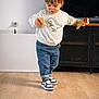 cabinet, casual_clothing, child, curious, dinosaur_print, flooring, home, indoor, jeans, playing, portrait, smiling, sneakers, standing, sweatshirt, television, toddler, toy, wooden_toy, young_child