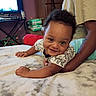 baby, infant, child, smiling, portrait, indoor, bedroom, bed, blanket, adult, support, curly_hair, brown_skin, short_sleeves, eyes, hands, television, dresser, furniture, cozy
