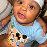 Kamari is registered to the contest to win money with this photo: baby, child, infant, portrait, indoors, sitting, supported, white_cap, blue_shirt, cartoon, mickey_mouse, disney, curious, open_mouth, large_eyes, smiling, adult_hands, family, bed, bedding