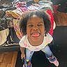 child, curly_hair, tongue_out, silly_face, standing, bed, blanket, stuffed_animal, rug, wooden_floor, clothing, jeans, socks, indoor, playful, happy, person, casual, young, fun