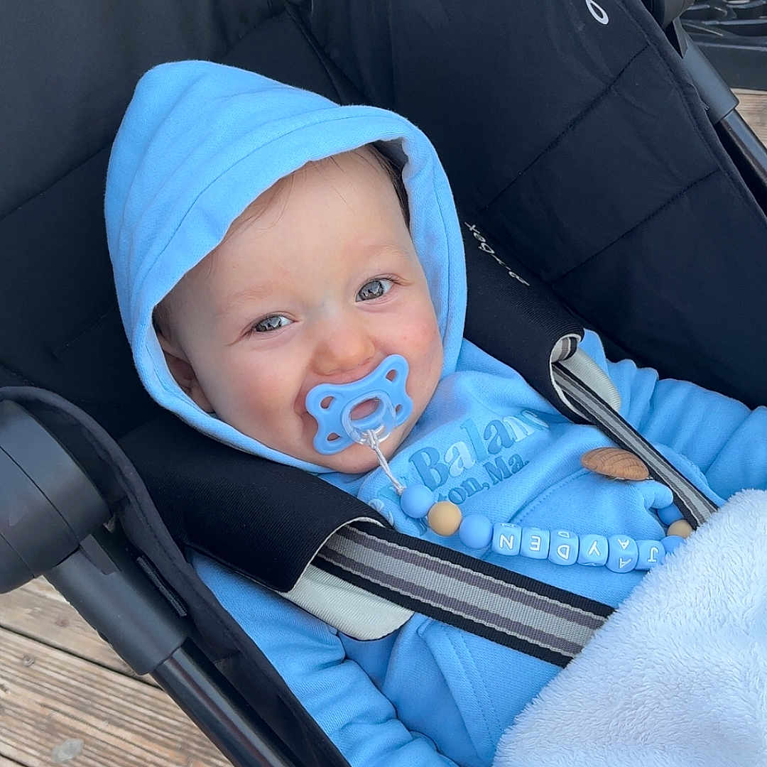 Jayden a rejoint le concours — aidez-le/la à gagner de superbes lots ! baby, beads, black_canopy, blanket, blue_clothing, cozy, eyes, face, hand, hood, hooded_jacket, infant, outdoor, pacifier, pacifier_clip, portrait, seat_belt, smile, stroller, wooden_deck
