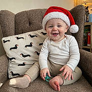 Archer is registered to the contest to win money with this photo: baby, child, smiling, santa_hat, pacifier, armchair, pillow, dachshund_pattern, cozy, indoor, lamp, furniture, cute, happy, festive, holiday, clothing, sitting, toddler, home