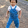 toddler, child, denim_overalls, white_shirt, white_sneakers, curly_hair, smiling, outdoor, gravel_path, sunlight, happy, walking, nature, greenery, casual_clothing, cute, portrait, daylight, person, fashion