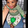 baby, balls, child, cute, face, green_clothing, grid_background, hand, happy, indoors, pajamas, person, playing, portrait, screen_capture, sitting, smiling, social_media, toddler, young_child