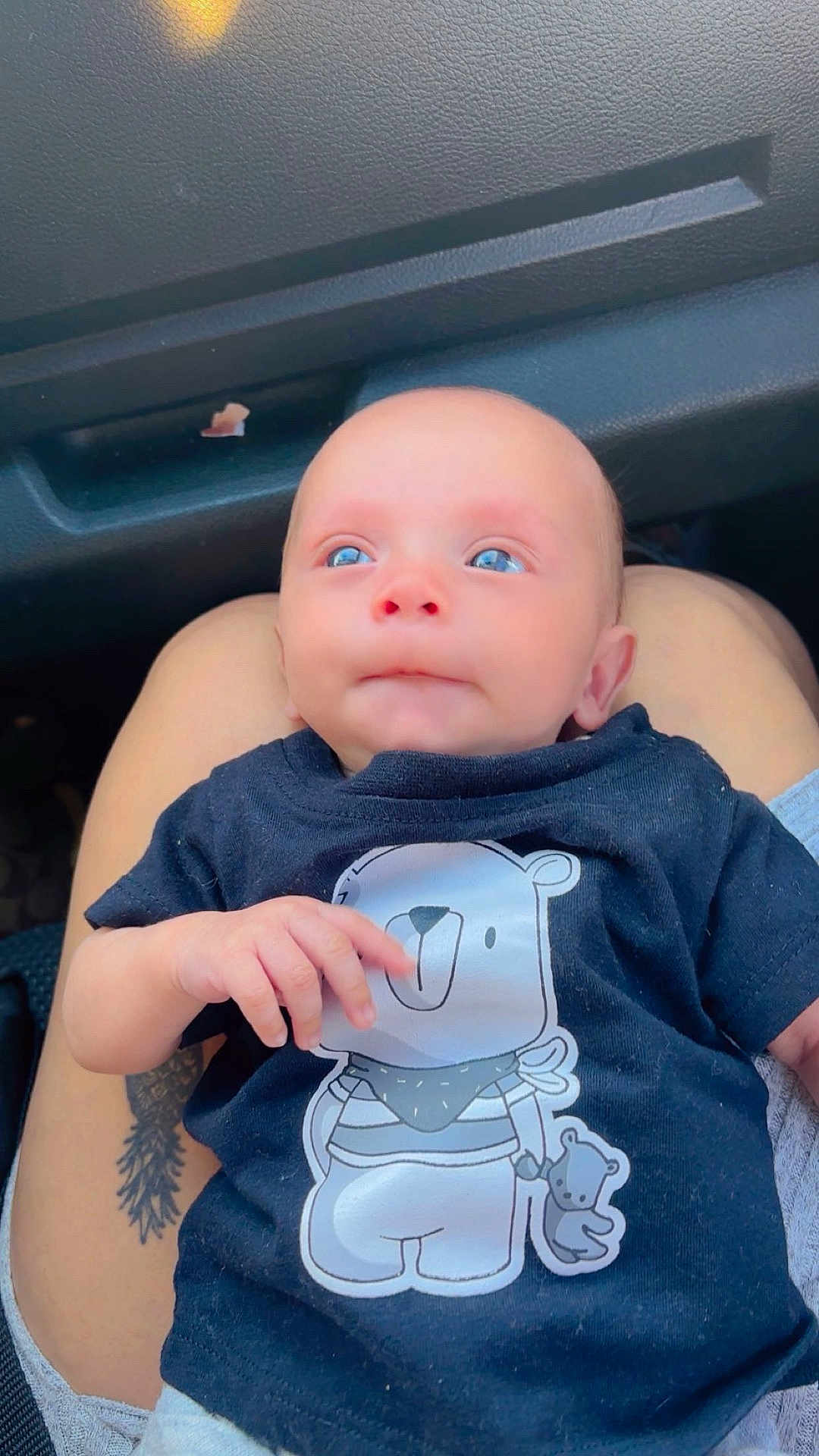Jorge is registered to the contest to win money with this photo: baby, infant, blue_eyes, tshirt, cartoon_bear, lap, person, car_interior, tattoo, hand, cute, child, portrait, casual_clothing, skin, head, sitting, expression, closeup, human