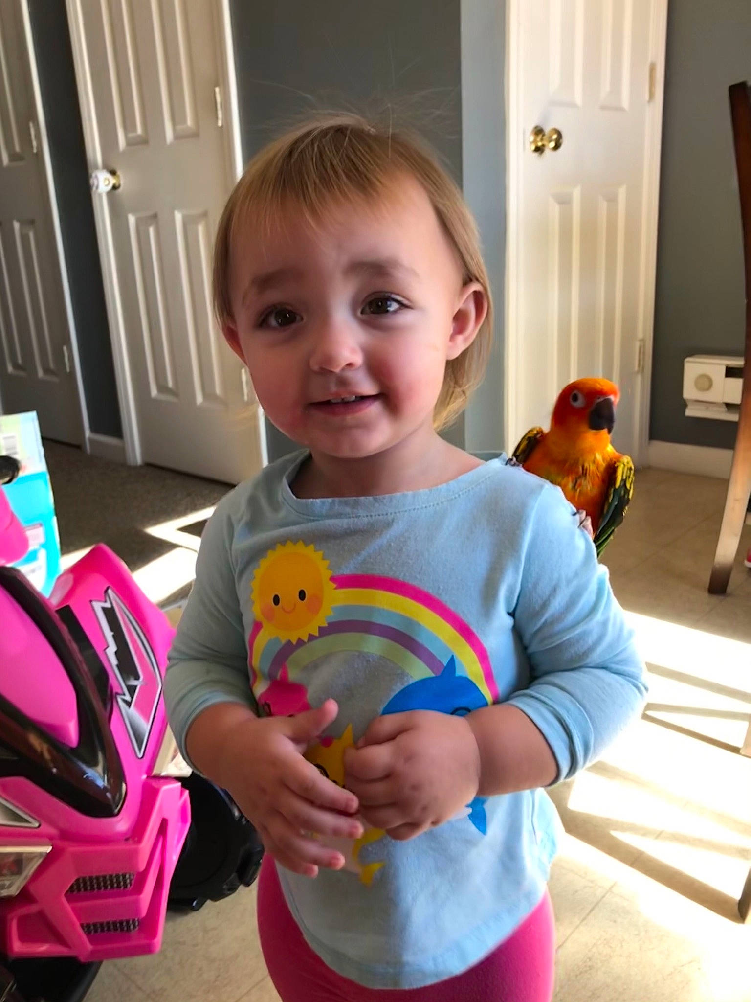 Ava is registered to the contest to win money with this photo: baby_toddler_clothing, balloon, child, door, finger, flooring, fun, happy, party_supply, person, riding_toy, room, smile, standing, stuffed_toy, t_shirt, tire, toddler, toy, wheel