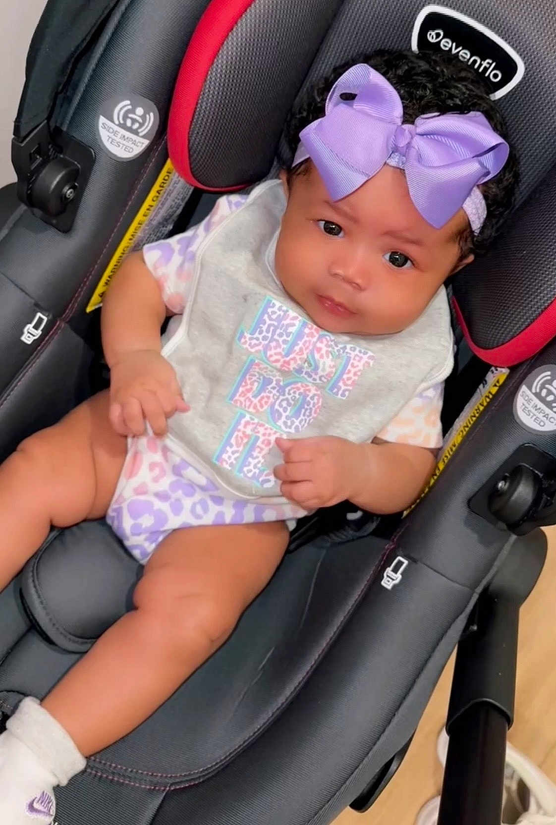 True is registered to the contest to win money with this photo: baby, car_seat, purple_bow, bib, clothing, infant, child, face, person, cute, sitting, indoor, footwear, nike, black_hair, skin, expression, looking, headband, legs