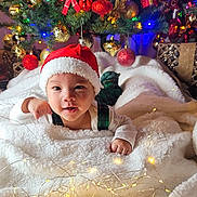Yujin is registered to the contest to win money with this photo: baby, child, santa_hat, christmas_tree, christmas_ornaments, fairy_lights, blanket, holiday, festive, smiling, cute, indoors, decorations, red, green, white, plaid, face, person, celebration