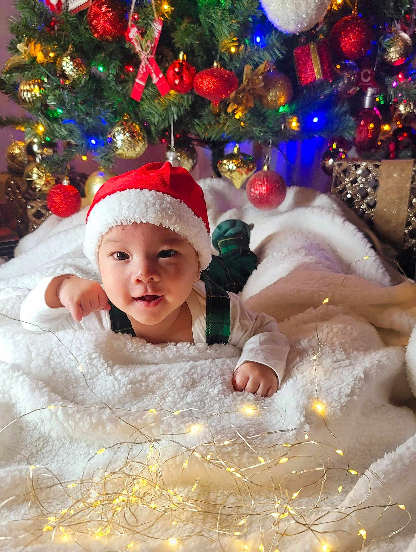 Yujin is registered to the contest to win money with this photo: baby, child, santa_hat, christmas_tree, christmas_ornaments, fairy_lights, blanket, holiday, festive, smiling, cute, indoors, decorations, red, green, white, plaid, face, person, celebration