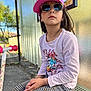 child, girl, sunglasses, cap, pink, bus_stop, bench, scooter, outdoor, sunny, patterned_pants, long_sleeve_shirt, cartoon_print, waiting, casual, daylight, metal_bench, trees, reflection, street