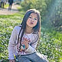 child, girl, sitting, daisies, grass, outdoor, sunlight, long_hair, casual_clothing, pointing, nature, greenery, plaid_pants, purple_shoes, portrait, daylight, expression, young, flower_field, park
