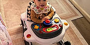 Mia is registered to the contest to win money with this photo: toddler, baby_walker, child, indoor, play, toy, carpet, curtain, furniture, floor, smiling, person, clothing, bib, pigtails, window, light, room, plastic, happy