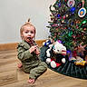 barefoot, child, chocolate, christmas_tree, cute, decorations, festive, floor, green_sweater, happy, holiday, holiday_season, indoors, plush_toys, pretzel, toddler, toy_bear, tree_lights, wood_floor, young_child