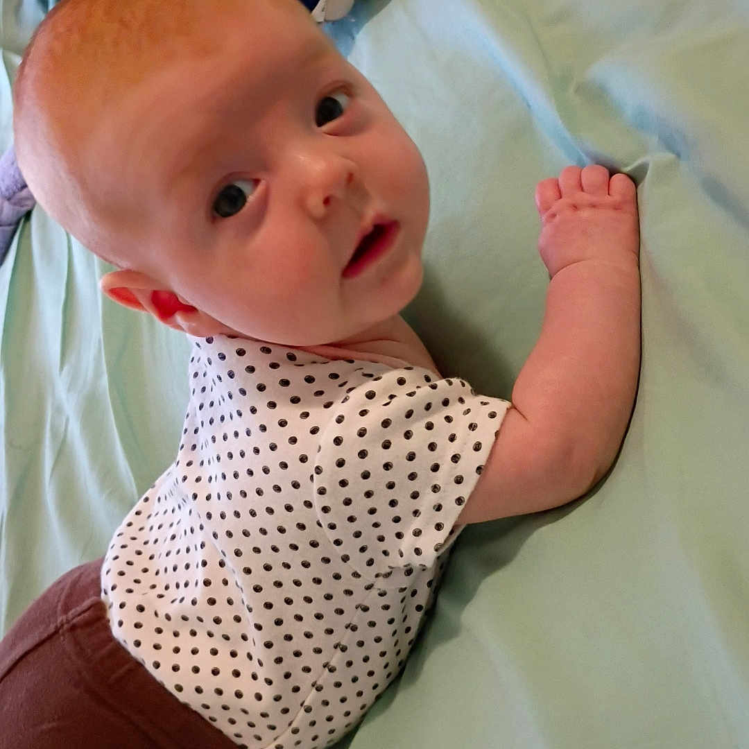 Eloise is registered to the contest to win money with this photo: baby, infant, polka_dot, shirt, brown_pants, bed, bedding, light_green, curious, face, child, person, lying_down, looking_back, indoor, soft_light, cute, small_hand, head, expression