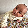 baby, infant, face, eyes, hand, onesie, blanket, floral_pattern, bed, wall, portrait, close_up, curious, lying_down, skin, gaze, newborn, room_interior, cozy, cute