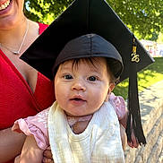 Raelynn joined the competition — help win amazing prizes! baby, graduation_cap, tassel, adult, smile, red_dress, outdoor, greenery, sunlight, tree, bib, pink_clothing, person, portrait, holding, face, happy, daylight, celebration, young_child