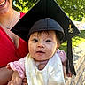 baby, graduation_cap, tassel, adult, smile, red_dress, outdoor, greenery, sunlight, tree, bib, pink_clothing, person, portrait, holding, face, happy, daylight, celebration, young_child