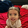 bed, blanket, child, clothing, cute, face, hair, happy, headboard, indoor, person, pigtails, pillow, portrait, red_bedspread, room, sitting, smiling, toddler, young_child