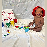 Kasyn joined the competition — help win amazing prizes! baby, child, smiling, red_hat, paint, paint_brush, white_sheet, colorful, messy, sitting, happy, indoor, playful, art, canvas, diaper, celebration, face, person, creative