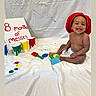 baby, child, smiling, red_hat, paint, paint_brush, white_sheet, colorful, messy, sitting, happy, indoor, playful, art, canvas, diaper, celebration, face, person, creative