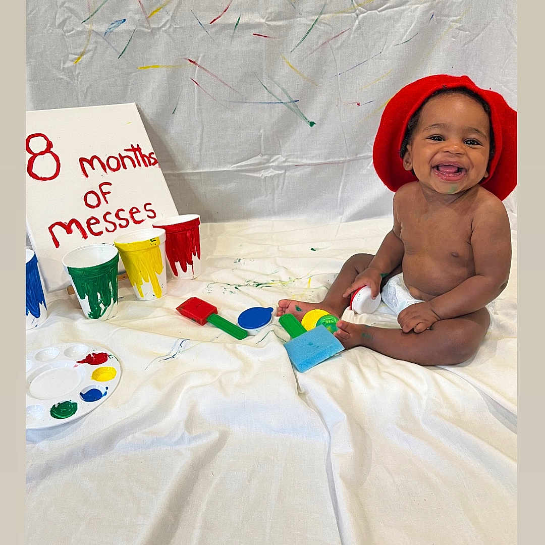 Kasyn joined the competition — help win amazing prizes! baby, child, smiling, red_hat, paint, paint_brush, white_sheet, colorful, messy, sitting, happy, indoor, playful, art, canvas, diaper, celebration, face, person, creative