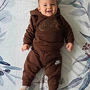 Mahé participe au concours pour gagner de l'argent avec cette photo : baby, child, smiling, brown_clothing, hoodie, pants, socks, lying_down, blanket, pattern, face, happy, infant, young_child, cute, indoor, person, portrait, apparel, footwear