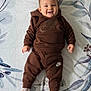 baby, child, smiling, brown_clothing, hoodie, pants, socks, lying_down, blanket, pattern, face, happy, infant, young_child, cute, indoor, person, portrait, apparel, footwear