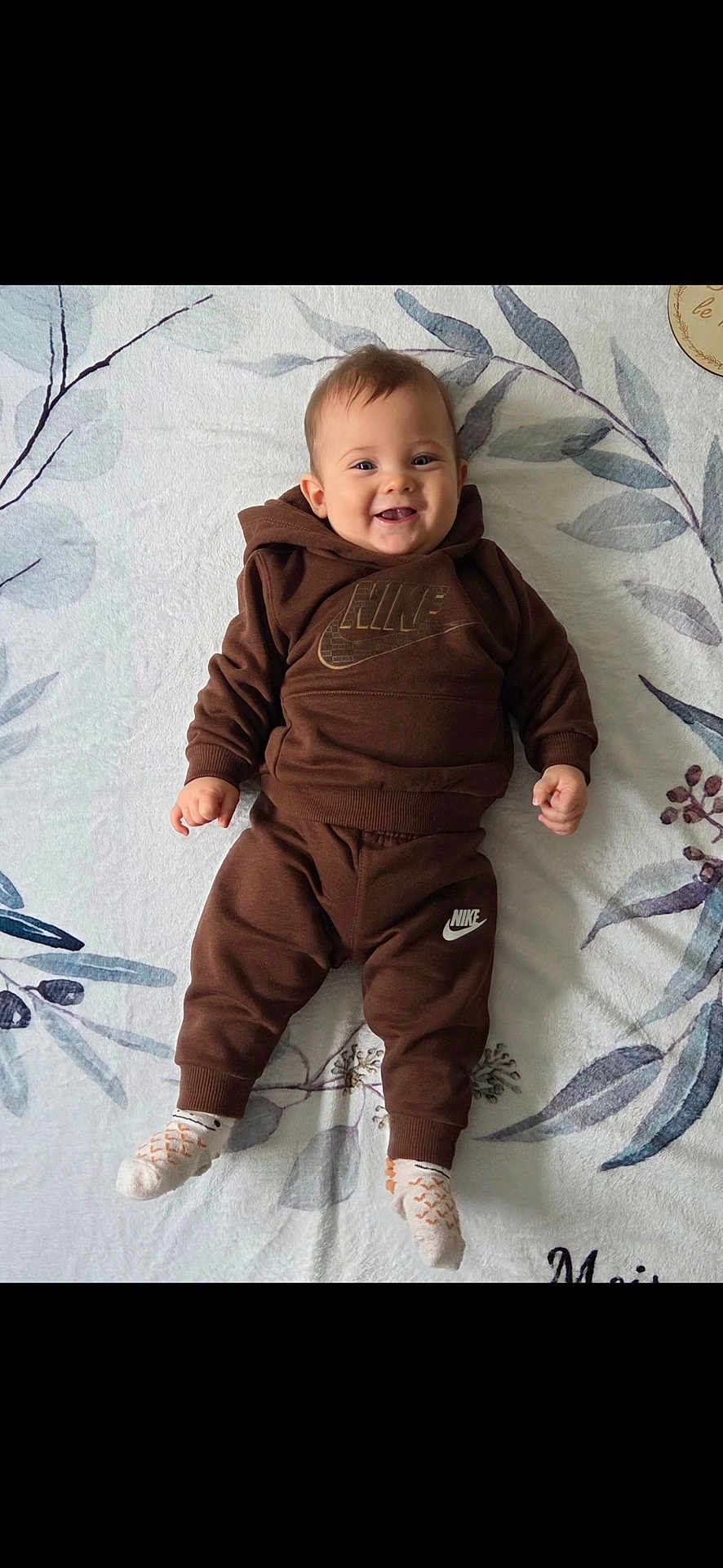 Mahé participe au concours pour gagner de l'argent avec cette photo : baby, child, smiling, brown_clothing, hoodie, pants, socks, lying_down, blanket, pattern, face, happy, infant, young_child, cute, indoor, person, portrait, apparel, footwear